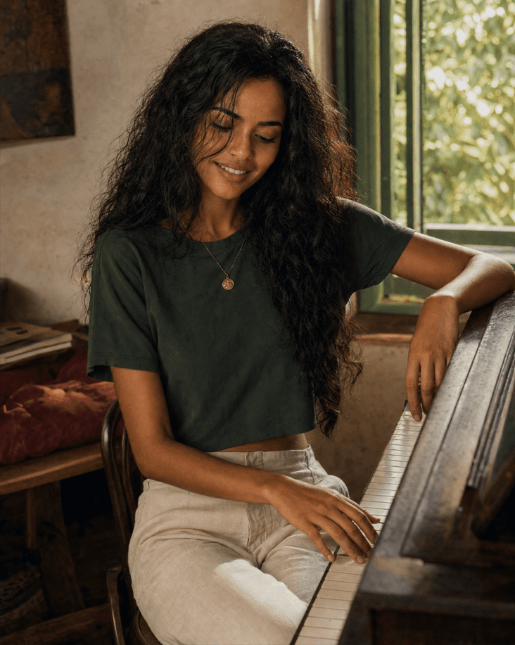 Vox Athena seated at an upright piano, smiling with her gaze lowered toward the keys.